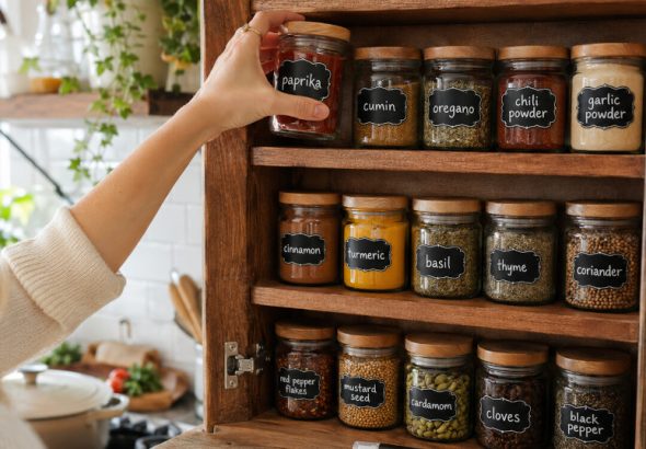 Organized kitchen spice cabinet with matching glass jars featuring handmade chalkboard labels, white chalk marker lettering, and a person reaching for spices while cooking in a bright, cozy kitchen.