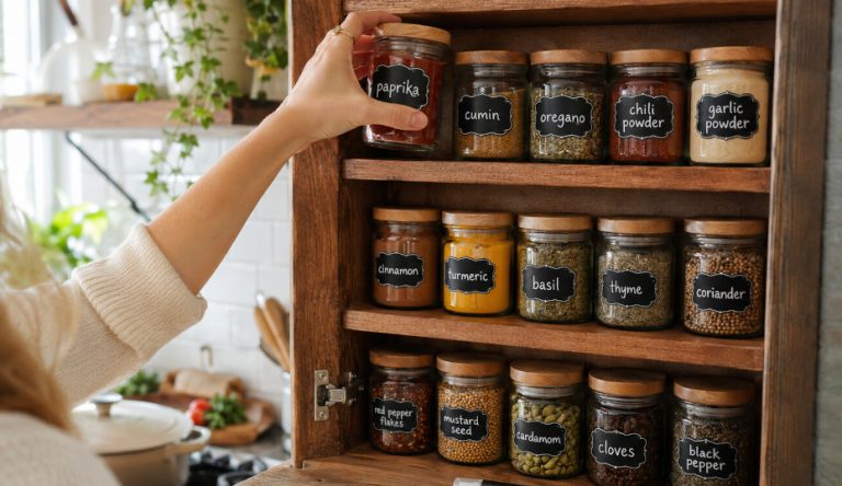 Organized kitchen spice cabinet with matching glass jars featuring handmade chalkboard labels, white chalk marker lettering, and a person reaching for spices while cooking in a bright, cozy kitchen.